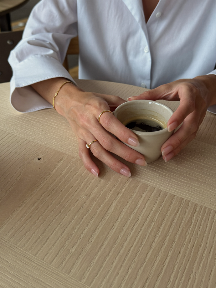 Hands wearing thin gold rings and a delicate gold bracelet, holding a ceramic coffee cup