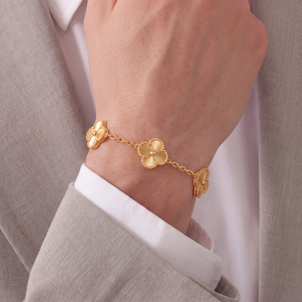 A gold chain bracelet with three clover-shaped links worn on a person's wrist, styled with a white dress shirt and a light gray blazer.