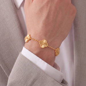 A gold chain bracelet with three clover-shaped links worn on a person's wrist, styled with a white dress shirt and a light gray blazer.